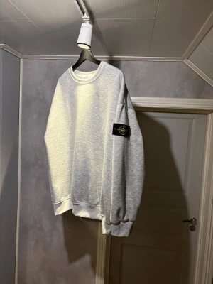 Grå sweatshirt från Stone Island - Snygg grå sweatshirt från Stone Island med klassisk rund hals och ribbade muddar. Ikonisk svart patch med gult och grönt Stone Island-märke på vänster ärm. Perfekt för en clean och avslappnad stil. Storlek XL, använd fåtal gånger, finns inga fula märken eller skav, ser ny ut. Pris går att diskuteras, skickas samma dag eller dagen efter köpet 