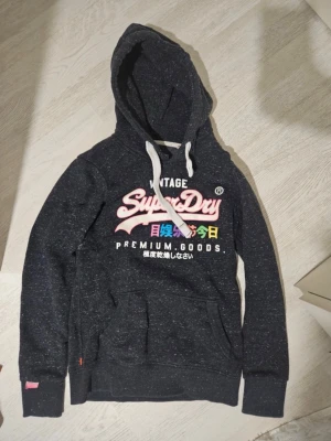 Mörkgrå Superdry hoodie med tryck - Säljer en mörkgrå hoodie från Superdry med färgglatt tryck på bröstet och japanska tecken. Hoodien har vit dragsko, känguruficka och ribbade muddar. Perfekt för dig som gillar streetwear och vill ha något bekvämt och snyggt.