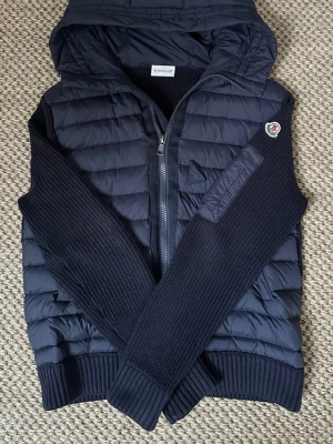 Mörkblå cardigan med huva från Moncler - Snygg mörkblå cardigan från Moncler med huva och dragkedja framtill. Västen har quiltad design, ribbade muddar och stickade detaljer på sidorna. Klassisk Moncler-logga på bröstet och en diskret patch. Perfekt för lager-på-lager och streetwear.