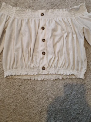 Vit offshoulder topp med knappar - Super söt vit offshoulder topp med långa ärmar och dekorativa knappar framtill. Toppen har smockdetaljer upptill och nedtill för en bekväm passform. 