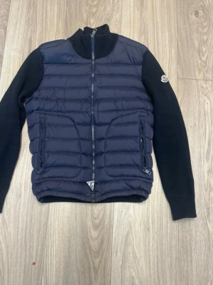  Moncler - Snygg mörkblå cardiganjacka från Moncler med quiltad front och stickade ärmar. Jackan har dragkedja framtill, två fickor med dragkedja och en Moncler-logga på ärmen. Perfekt för lager-på-lager och en stilren look.