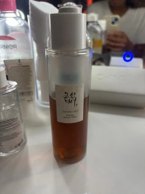 Ginseng Essence Water toner - En genomskinlig plastflaska med vit kork innehållande Ginseng Essence Water toner från Beauty of Joseon. Tonern har en brunaktig färg och är perfekt för att återfukta och fräscha upp huden. Flaskan har en stilren och minimalistisk design med etikett på koreanska.