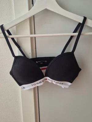 Svart bh från Tommy Hilfiger - Snygg svart bh från Tommy Hilfiger med tunna axelband. Under bysten finns en vit resår med logotyp och klassiska ränder i rött, vitt och blått. Perfekt för dig som gillar sportig och stilren design.