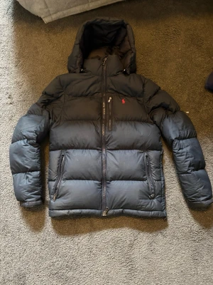 Svart dunjacka med huva från Polo Ralph Lauren - Snygg svart dunjacka från Polo Ralph Lauren med huva och dragkedja framtill. Jackan har två stora fickor med dragkedja och en bröstficka. Klassisk röd logga på bröstet och quiltad design som håller värmen under kalla dagar.
