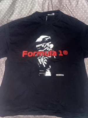 Svart Formula 1 t-shirt Divided - Baggy Cool svart t-shirt från Divided med stort Formula 1-tryck i rött och vitt framtill. Motiv av en racerförare och texten 'Live to race' ger en sportig vibe. Klassisk rund hals och korta ärmar. Perfekt för dig som älskar motorsport.