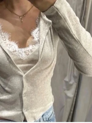 Beige omlottopp med spetsdetaljer - Snygg beige omlottopp med långa ärmar och v-ringning. Under toppen syns en matchande linnetopp med vit spetskant som ger en extra feminin touch. Perfekt att styla med jeans för en trendig look.