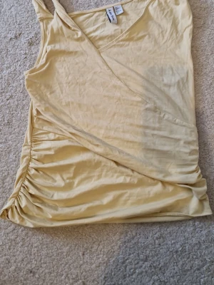 Ljusgul topp - Säljer en ljusgul omlottopp med smala axelband och snygga rynkdetaljer på sidan. Toppen har en mjuk och stretchig känsla.. Materialet är en bomullsblandning. Enkel men trendig design.