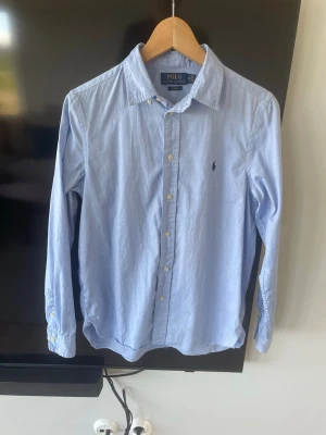Ljusblå skjorta från Polo Ralph Lauren - Klassisk ljusblå skjorta från Polo Ralph Lauren med knappstängning framtill och broderad logga på bröstet. Skjortan har lång ärm, normal passform och är tillverkad i bomull. Perfekt för en clean och stilren look. Liten fläck/missfärgning som ni kan se på tredje bilden men ingenting man ser när skjortan är på. Tveka inte på att ställa frågor!