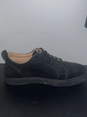 Loubs, svarta sneakers med röd sula - Stilrena svarta sneakers i mocka med lågt skaft och svarta skosnören. Skorna har en unik röd sula med grafiskt mönster undertill och beige insida. Perfekta för dig som vill ha en clean look med en twist.