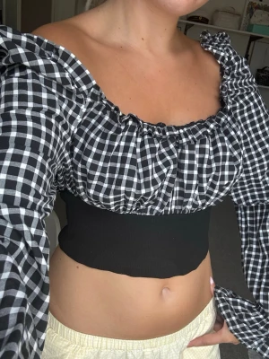 Svartvitrutig croppad blus - Trendig croppad blus med svartvitt rutigt mönster och långa puffärmar. Toppen har en bred, ribbad midja i svart och en offshoulder-skärning som ger en snygg siluett. Perfekt för dig som vill sticka ut med en cool och ungdomlig stil.