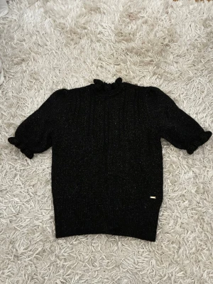 Svart stickad topp med glitter - Snygg svart stickad topp med diskret glittereffekt. Modellen har puffärmar, volangkant vid halsen och kortare ärmar med rynkade avslut. Perfekt för dig som vill ha något extra till din outfit. Tyvärr har två diamanter lossnat i knappen, men det är varken något som syns eller som man tänker på 