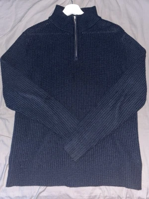 Mörkblå stickad halvzip tröja - Mörkblå stickad tröja med halv dragkedja och hög krage. Tröjan har lång ärm och ribbstickad struktur som ger en stilren look. Perfekt för lager på lager och passar till både jeans och byxor.