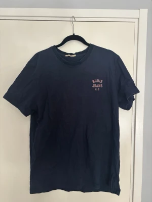 Nudie jeans t-shirt - Snygg mörkblå t-shirt från Nudie Jeans med tryck på bröstet. Klassisk passform och rund halsringning. Tillverkad i mjuk bomull som känns skön mot huden. Perfekt till jeans eller shorts för en enkel och clean look.