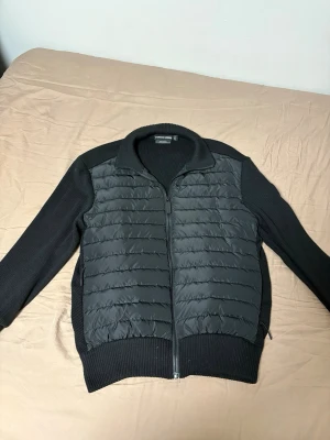 Svart quiltad cardigan med dragkedja - Snygg svart cardiganjacka med quiltad framsida och stickade ärmar samt rygg. Jackan har dragkedja framtill, ribbade muddar och hög krage. Perfekt för lager-på-lager och ger en sportig look.