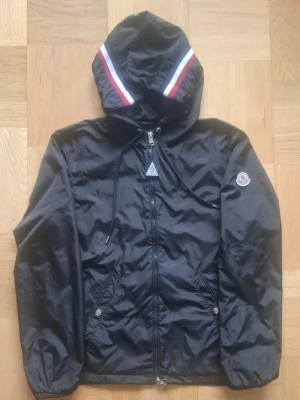 Svart Moncler vindjacka - Snygg svart vindjacka från Moncler med dragkedja framtill, två fickor med tryckknappar och justerbar huva med vita och röda ränder. Jackan har Moncler-logga på ärmen och elastiska muddar vid ärmsluten. Perfekt för blåsiga dagar.