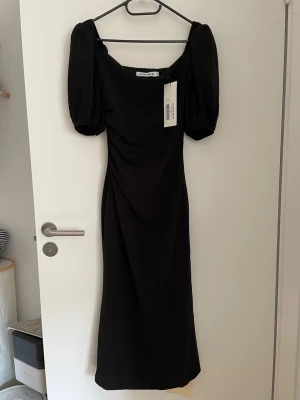 ADOORE vellore dress stl 32 - Elegant svart långklänning med puffiga kortärmar och rak passform. Klänningen har en fyrkantig ringning och är tillverkad i ett slätt tyg som ger ett stilrent intryck. Perfekt för dig som vill ha en klassisk och trendig look.