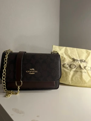 Mörkbrun Mini Klare från Coach - Snygg mörkbrun handväska från Coach med klassiskt monogrammönster och guldfärgad kedja. Väskan har ett lock med magnetlås och detaljer i mörkbrunt läder. Perfekt storlek för det viktigaste och ger en lyxig känsla till din outfit.