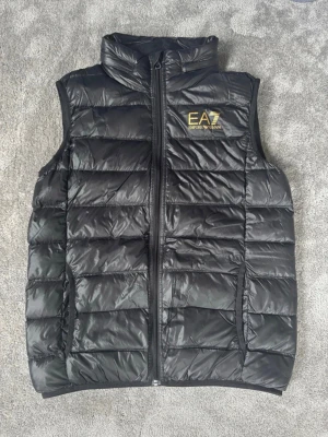 EA7 Emporio Armani- svart/guld - Jag säljer en väldigt snygg svart väst från Emporio Armani, som aldrig har används, alltså är den som att köpa en ny! Rekommenderar verkligen att köpa med tanke på vad den egentligen kostar som ny! Västen passar både tjejer och killar! Storlek: 160 cm (14 år)