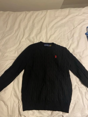 Svart kabelstickad tröja Polo Ralph Lauren - Kabelstickad svart tröja från Polo Ralph Lauren med klassisk röd logga broderad på bröstet. Tröjan har rund halsringning och ribbade muddar vid ärmslut och nederkant. Perfekt för dig som gillar stilrena och tidlösa plagg. Den är L med den passar S/M