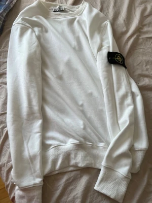  Stone Island - Säljer en clean vit sweatshirt från Stone Island med den klassiska svarta loggan på vänster ärm. Tröjan har rund hals, ribbade muddar och är tillverkad i mjukt bomullsmaterial. Perfekt för dig som gillar streetwear och enkel stil.passar som L också