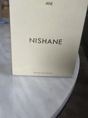 Nishane Ani Extrait de Parfum 50ml - Nishane Ani Extrait de Parfum i en stilren beige kartong. Flaskan innehåller 50 ml exklusiv parfym med intensiv doft, perfekt för dig som vill sticka ut. Förpackningen har minimalistisk design med tydlig logotyp och detaljer i svart text.