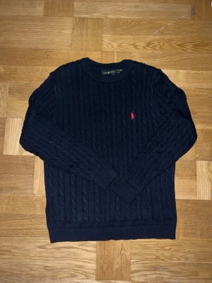 Mörkblå kabelstickad tröja Ralph Lauren - Kabelstickad mörkblå tröja från Ralph Lauren med rund halsringning och den klassiska röda logotypen broderad på bröstet. Tröjan har långa ärmar och ribbade muddar vid ärmslut och nederkant. Storlek M men passar även stor S. Perfekt för dig som gillar tidlös och stilren design.