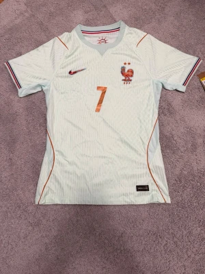 Vit Frankrike fotbollströja Nike - Snygg vit fotbollströja från Nike med Frankrikes landslagsemblem och nummer 7 på bröstet. Tröjan har röda och blå detaljer, korta ärmar och V-ringad krage med randig insida. Perfekt för dig som älskar fotboll och vill stötta Les Bleus!