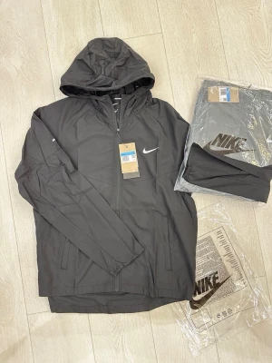Svart vindjacka från Nike med huva - Snygg svart vindjacka från Nike med dragkedja, justerbar huva och två sidofickor. Jackan har en vit Nike-logga på bröstet och är tillverkad i lätt, vindtätt material. Perfekt för blåsiga dagar och enkel att matcha med olika outfits.