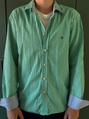 Morris skjorta - Snygg grön och vit rutig skjorta med klassisk krage och långa ärmar. Skjortan har knappar framtill och en liten broderad detalj på bröstet. Perfekt för en avslappnad men stilren look.