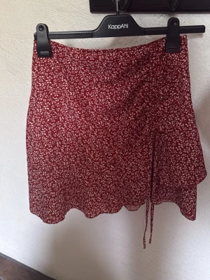 Röd blommig omlottkjol med knyt - Söt röd omlottkjol från KappAhl med vitt blommönster och knyt i sidan. Kjolen har en vågig nederkant och är perfekt för varma dagar. Lätt och luftig modell som ger en avslappnad känsla.