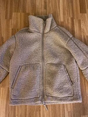 Beige teddyjacka från H&M - Mysig beige teddyjacka från H&M med hög krage och dragkedja framtill. Jackan har två stora fickor med snedställda öppningar och är gjord i fluffigt teddy-material som håller dig varm. Perfekt för kyliga dagar och enkel att matcha med olika outfits.