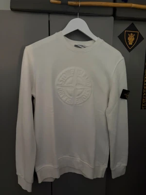 Vit sweatshirt från Stone Island - Snygg vit sweatshirt från Stone Island med det klassiska broderade logotypmärket på ärmen och ett stort präglat Stone Island-emblem på bröstet. Tröjan har rund hals, långa ärmar och ribbade muddar. Perfekt för en clean och stilren look.