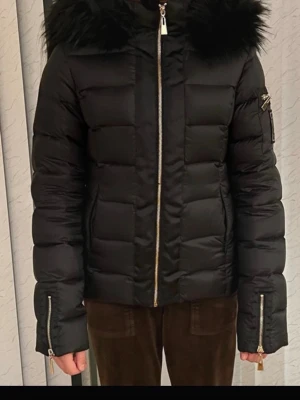 Svart dunjacka med fuskpäls - Snygg svart dunjacka med stor huva och fuskpälsdetalj. Jackan har quiltad design, dragkedja framtill och dragkedjor vid ärmsluten. Ficka med dragkedja på ena ärmen ger extra stilpoäng. Perfekt för kalla vinterdagar.