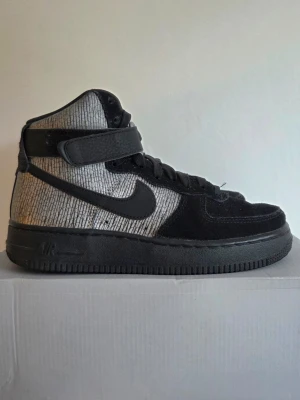 Nike Air Force 1 High svart/silver - Nike Air Force 1 High sneakers i svart och silver med högt skaft och kardborreband runt ankeln. Ovandelen har ett unikt mönster i silver och svart mocka på tå och snörning. Klassisk svart sula och Nike Swoosh på sidan. Perfekt för dig som gillar streetwear.