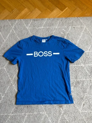 Blå BOSS t-shirt med tryck - Snygg blå t-shirt från BOSS med vit logga tryckt framtill. Klassisk rund halsringning och korta ärmar. Tillverkad i mjuk bomull som känns skön mot huden. Perfekt för en avslappnad och stilren look.