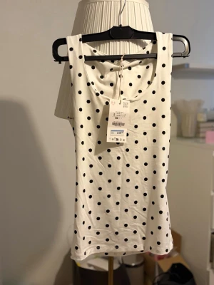 Vit prickig linnetopp - Snygg vit linnetopp med svarta prickar och rund halsringning. Perfekt för dig som gillar en enkel men lekfull stil. Toppen är ärmlös och har en rak passform som passar till det mesta.