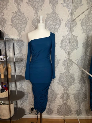 Blå one shoulder långklänning - Trendig blå långklänning med one shoulder-design och långa ärmar. Klänningen har rynkade detaljer längs sidan och nedtill, vilket ger en snygg siluett. Perfekt för dig som vill sticka ut med en unik och modern look.