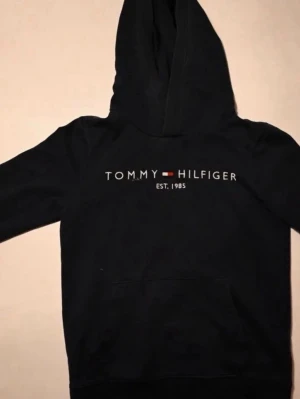 Svart hoodie från Tommy Hilfiger 152-164 - Snygg svart hoodie från Tommy Hilfiger med broderad logga och text 'EST. 1985' på bröstet. Klassisk huva och magficka framtill. Perfekt för en chill och stilren look.