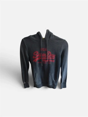 Mörkgrå Superdry hoodie med tryck - Snygg mörkgrå hoodie från Superdry med röd logga och japansk text på bröstet. Klassisk känguruficka framtill och justerbar huva med dragsko. Tillverkad i mjukt bomullsmaterial som är skönt mot huden. Perfekt för en avslappnad streetwear-look.