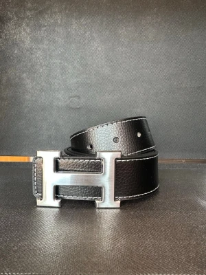 Svart skärp med stor H-spänne - Snyggt svart bälte i skinn med ett stort, silverfärgat H-format spänne. Bältet har vit kontrastsöm längs kanterna och en stilren, klassisk look som passar till många outfits.