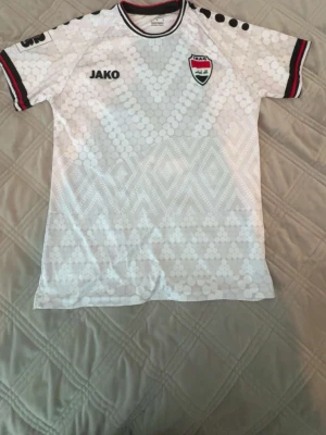 Vit Irak fotbollströja från Jako - Snygg vit fotbollströja från Jako med Irak-emblem på bröstet. Tröjan har ett geometriskt mönster, svarta och röda detaljer på ärmsluten samt svarta prickar på axlarna. Perfekt för dig som vill stötta Irak på match eller träning.