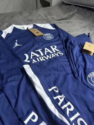 Blå PSG träningsset med vita detaljer - Säljer ett blått PSG träningsset med vita detaljer och logga på både tröja och byxor. Setet har dragkedja vid halsen, tryckt Qatar Airways sponsor och Jordan-logga. Materialet är lätt och andas, perfekt för fotbollsträning eller gym. Om du vill inte köpa hela set så kan du skriva till oss och tröjans priset är 300kr och byxorna 400kr