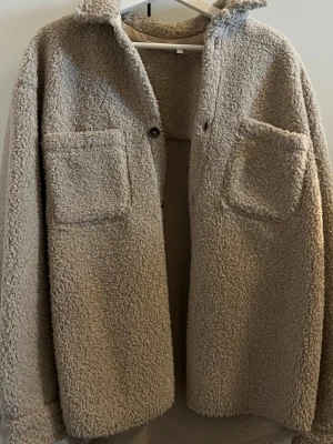 Beige oversize teddyjacka med fickor - Mysig beige teddyjacka med två stora bröstfickor och krage. Jackan har en oversized passform och stängs med knappar framtill. Perfekt för att hålla värmen under kyliga dagar och ger en trendig look.