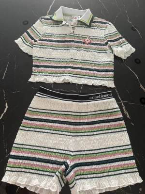 Randigt set från Casablanca - Säljer ett tvådelat set från Casablanca med kortärmad topp och shorts i smockat material. Setet är vitt med tunna ränder i grönt, rosa och mörkblått. Toppen har krage och knapp, shortsen har elastisk midja med logga. Båda delarna har volangkant. 100% bomull