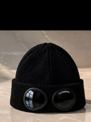 Svart mössa med goggles från C.P. Company - Säljer en svart ribbstickad mössa från C.P. Company med ikoniska goggles framtill. Mössan har uppvikt kant och är tillverkad i ett mjukt, värmande material. Perfekt för dig som vill sticka ut med en streetwear-inspirerad look.