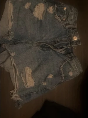 Blå jeansshorts - Säljer ett par blå denimshorts med slitna detaljer och fransiga kanter. Shortsen har hög midja, klassiska fem fickor och dragkedja framtill. Perfekta för en avslappnad och trendig look i sommar.