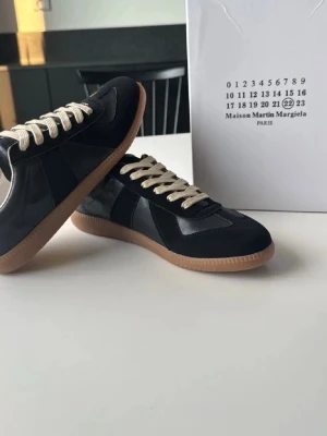 Maison Margiela svarta sneakers - Svarta sneakers från Maison Martin Margiela med beige snörning och klassisk gummisula. Skorna har en stilren design med inslag av mocka och läder, perfekt för dig som gillar minimalistisk och trendig stil.