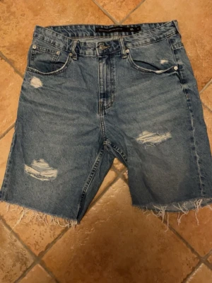 Blå denimshorts med slitningar - Säljer ett par blå jeansshorts i klassisk denim med slitna detaljer och råa kanter. Shortsen har fem fickor, knappgylf och en relaxed passform. Perfekta för sommaren och ger en avslappnad vibe.