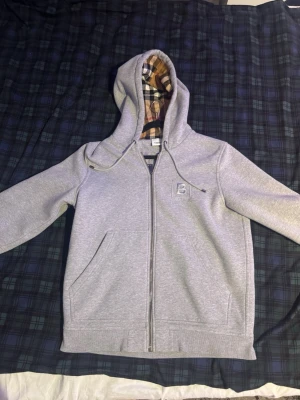 Grå zip hoodie från Burberry - Snygg grå zip hoodie från Burberry med broderad logga på bröstet och klassiskt rutigt foder i huvan. Hoodien har dragsko i huvan, två fickor fram och ribbade muddar. Perfekt för en chill och stilren look.