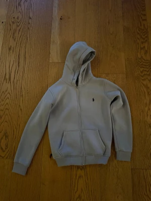  Beige hoodie med dragkedja - Snygg beige hoodie med dragkedja och huva. Tröjan har en klassisk känguruficka framtill och ribbade muddar vid ärmar och nederkant. Liten broderad logga på bröstet ger en stilren touch. Perfekt för en avslappnad och trendig look.
